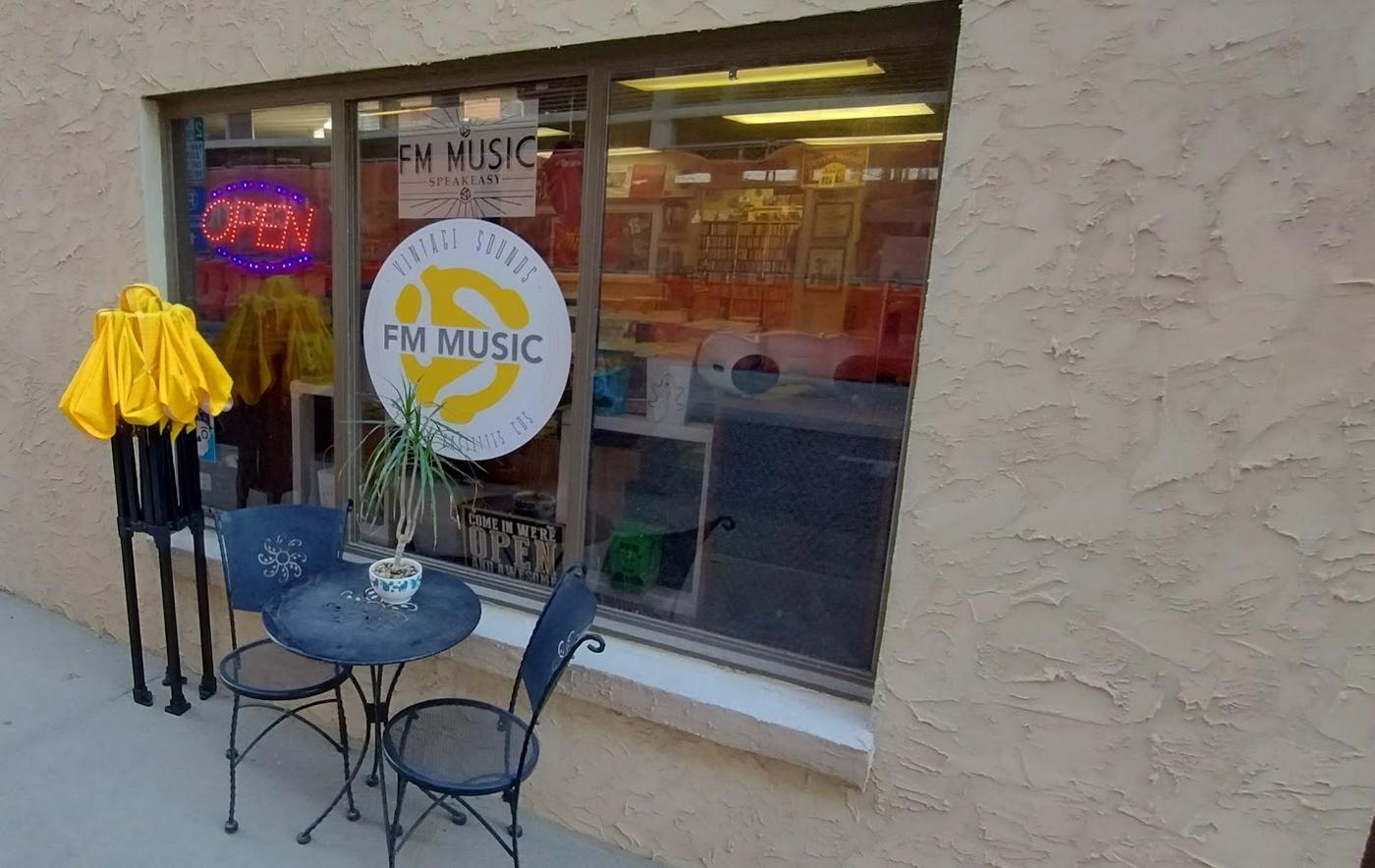 The shop front from the sidewalk: the window roundel, a neon open sign and a bistro table under a yellow umbrella.